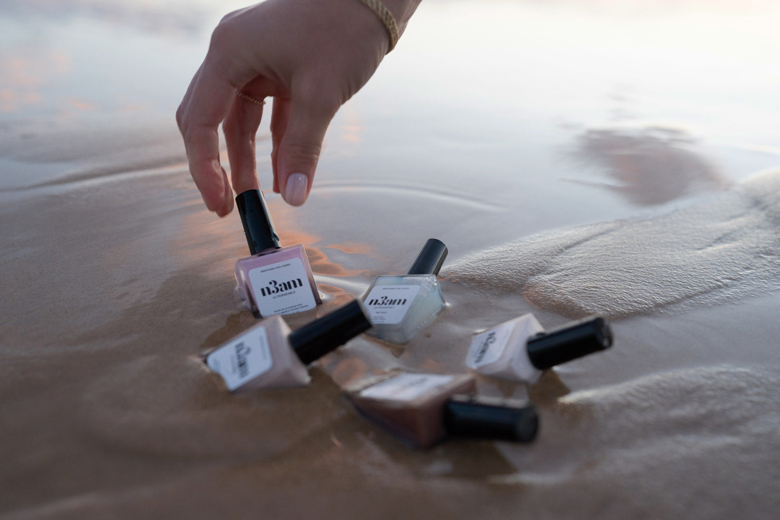 Main d'une femme portant du vernis perméable rose qui attrape un vernis à ongles posé sur le sable humide de la mer avec d'autre vernis de la marque n3am